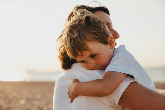 Elternteil hält sein Kind im Arm am Strand bei Sonnenuntergang, ruhiger Moment zwischen Eltern und Kita-Kind.